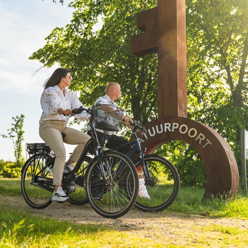 Man en vrouw op fiets in landschap met grote sleutel met daarop de tekst Natuurpoort