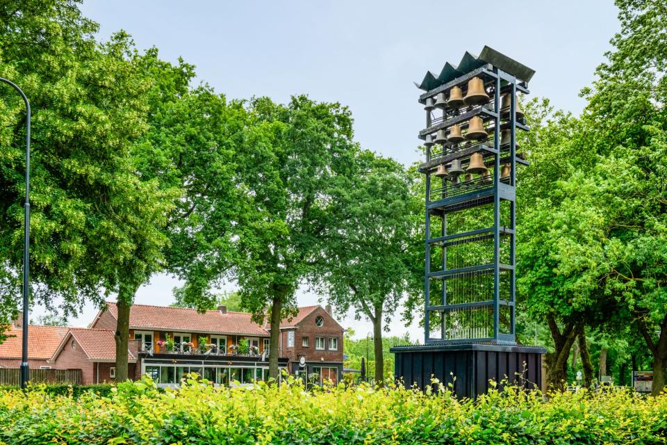 Struiken, bomen en toren met klokken met op de achtergrond een gebouw met schuin dak