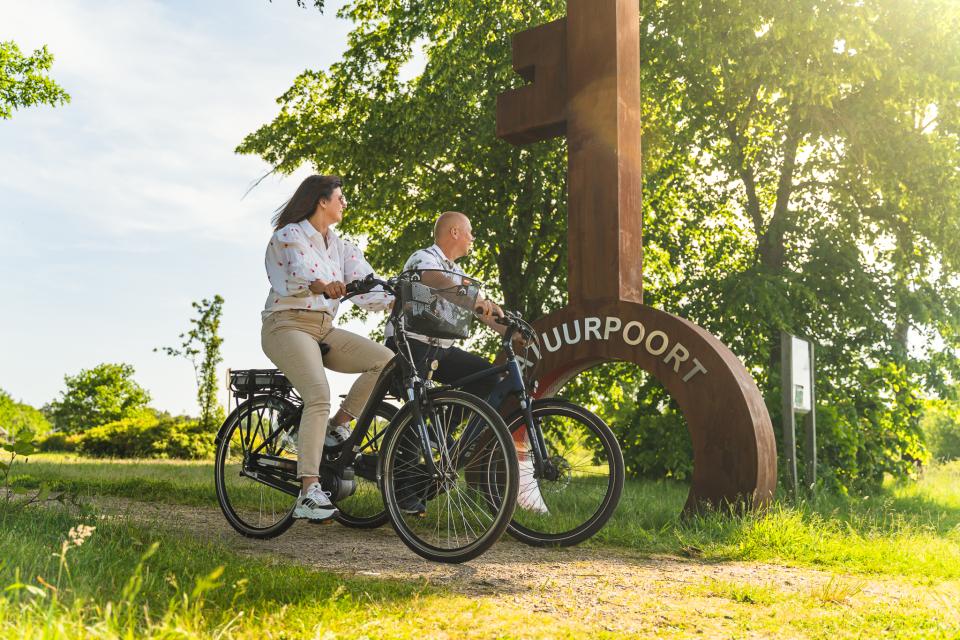 Man en vrouw op fiets in landschap met grote sleutel met daarop de tekst Natuurpoort