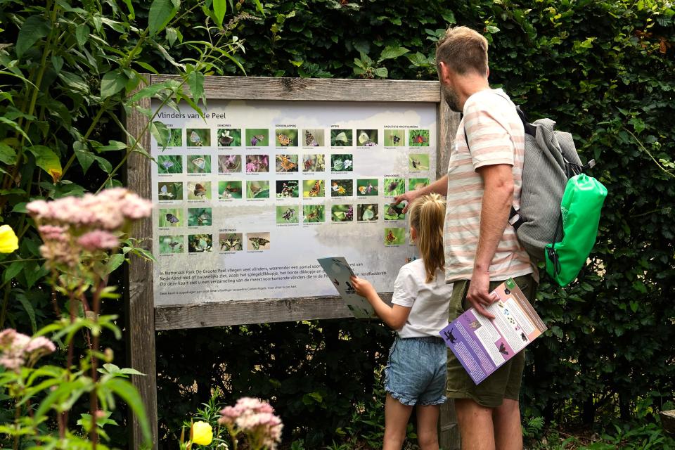 Man en meisje staan tussen planten en wijzen naar bord met plaatjes van vlinders